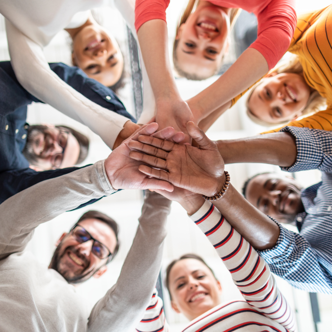 A group of people putting their hands in to form a team. The perspective is taken from the floor, facing upwards. The people are smiling.